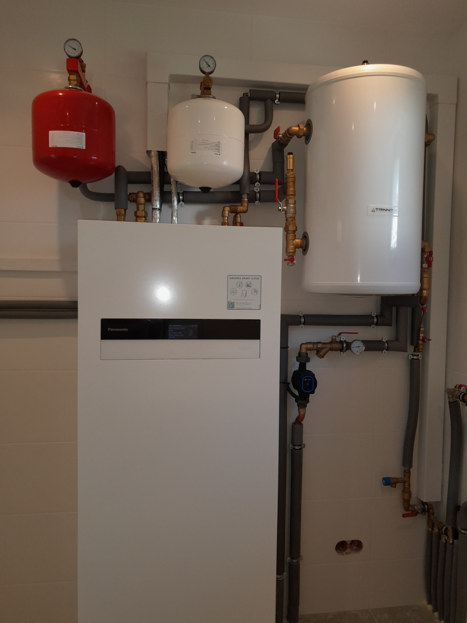 Biała pompa ciepła Panasonic Aquarea Smart Cloud z czerwonym i białym zbiornikiem wyrównawczym oraz białym zasobnikiem ciepłej wody marki Trinnity, widoczne rury i zawory, instalacja...