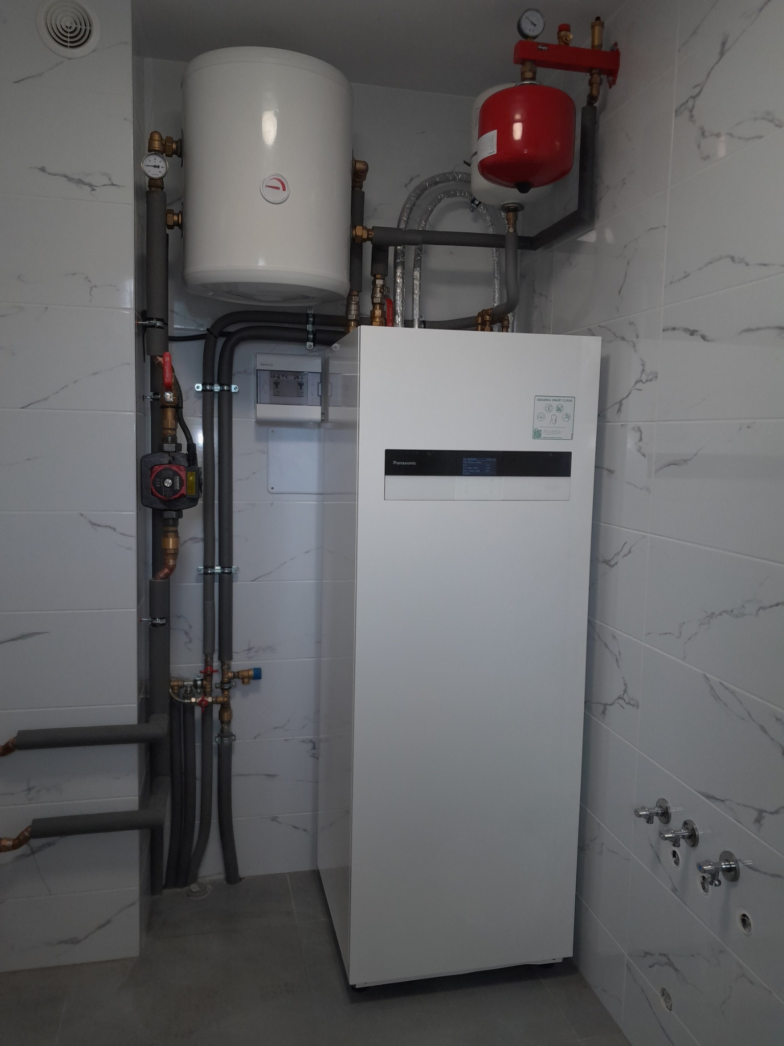Biała pompa ciepła Panasonic Aquarea Smart Cloud z czerwonym zbiornikiem wyrównawczym i białym zasobnikiem ciepłej wody, rury instalacji grzewczej z izolacją, zamontowane na tle białych płytek...