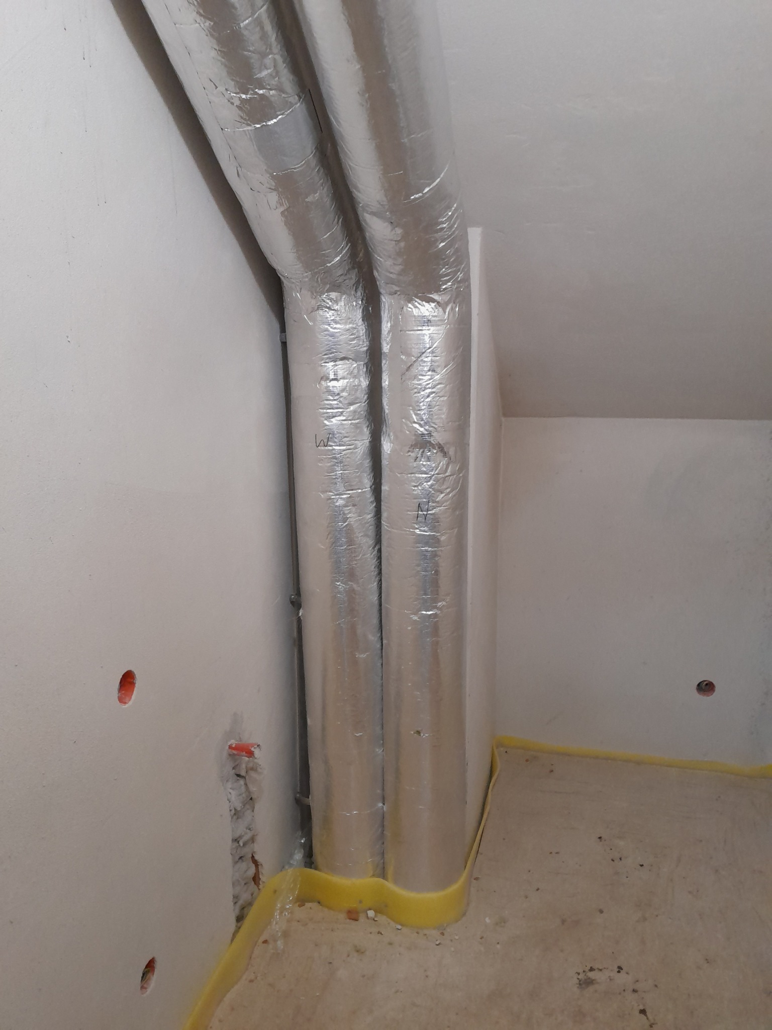 Dwie rury w srebrnej izolacji termicznej, z widocznymi napisami 'W' i 'N', prowadzone wzdłuż ściany, zabezpieczone żółtą taśmą malarską przy podłodze w trakcie prac wykończeniowych.