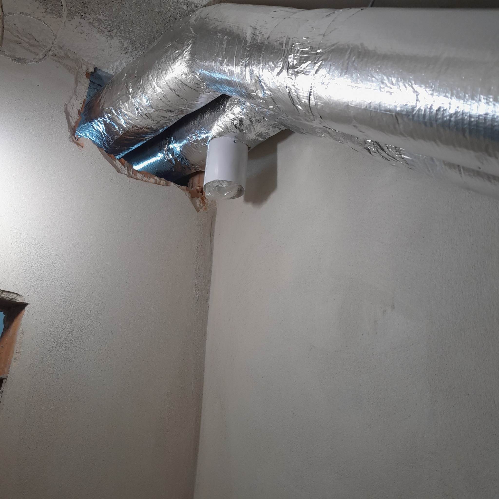 Fragment instalacji wentylacyjnej z aluminiową izolacją, widoczny biały element łączący rury, tło stanowią szare ściany i fragment sufitu z widoczną izolacją.
