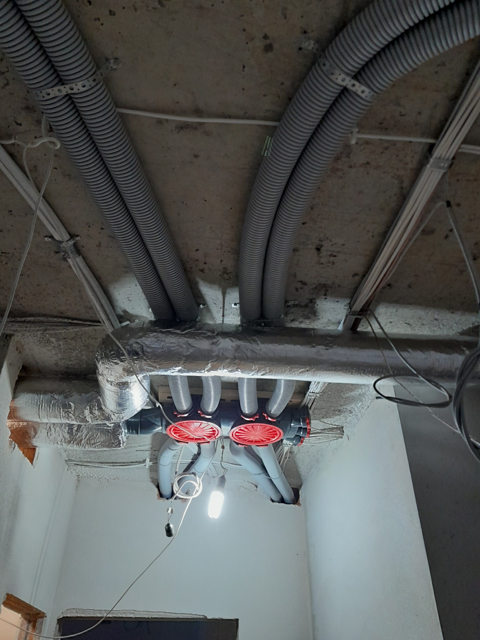 Widok z dołu na sufit z zamontowaną instalacją wentylacyjną: elastyczne rury wentylacyjne przymocowane do betonowego sufitu, rozdzielacz z czerwonymi elementami, przewody izolowane folią aluminiową...