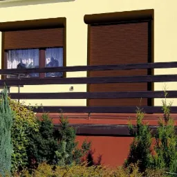 Brązowe rolety zewnętrzne na balkonie domu z żółtą elewacją, widoczne rośliny w ogrodzie.