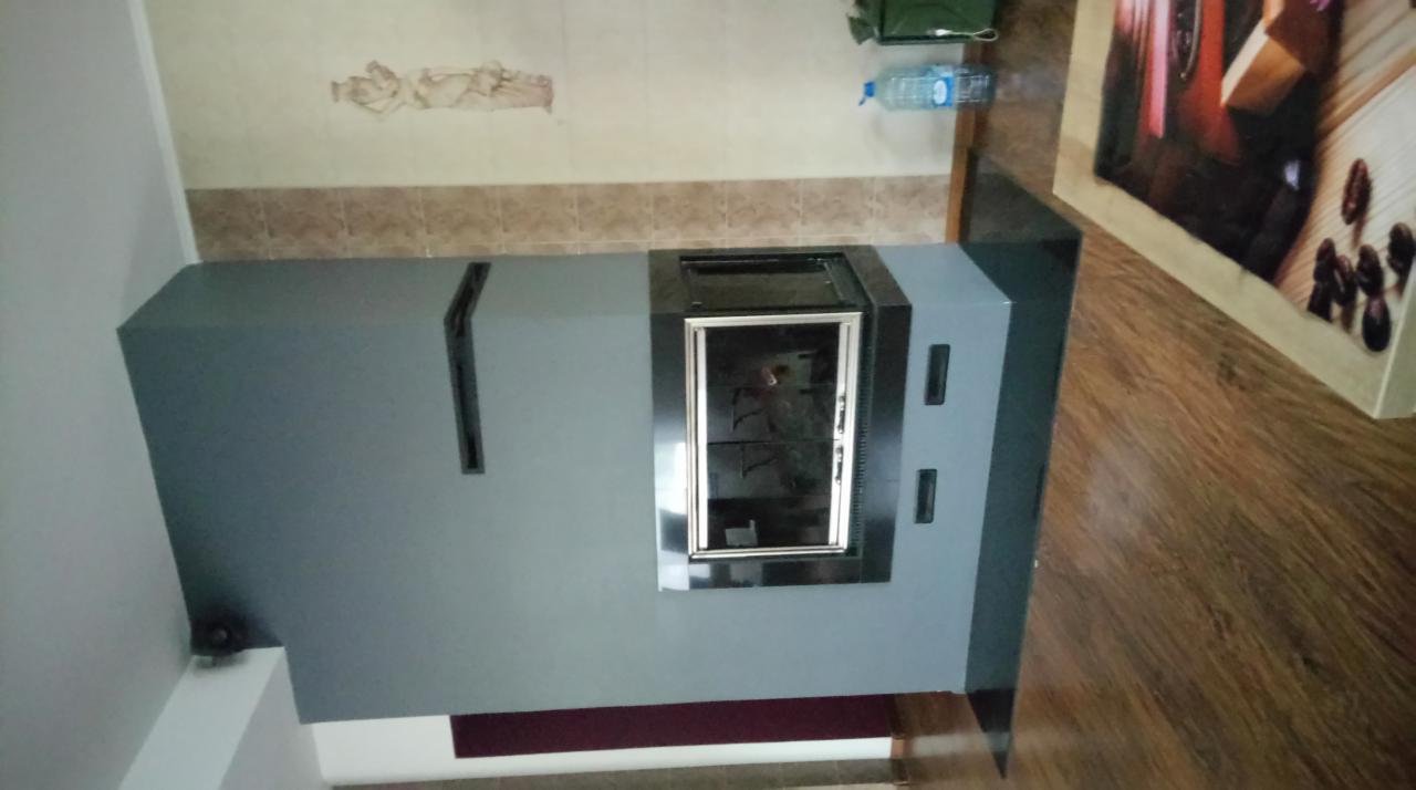 Szary kominek obudowany płytą karton gips z metalowym wkładem kominkowym, widoczny fragment ściany z dekoracyjnym elementem i brązowymi płytkami, drewnopodobna podłoga.