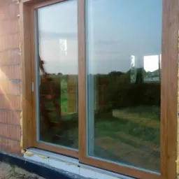 Montaż dużych, brązowych drzwi przesuwnych na surowej ścianie z cegły, widoczne pianka montażowa i teren budowy.