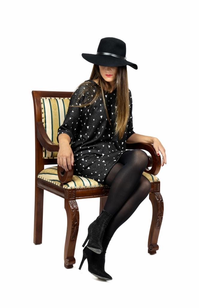 Kobieta w czarnym kapeluszu i sukience w trójkąty siedzi na antycznym krześle z tapicerowanym siedziskiem i oparciem w paski, ma na sobie czarne rajstopy i botki na obcasie.