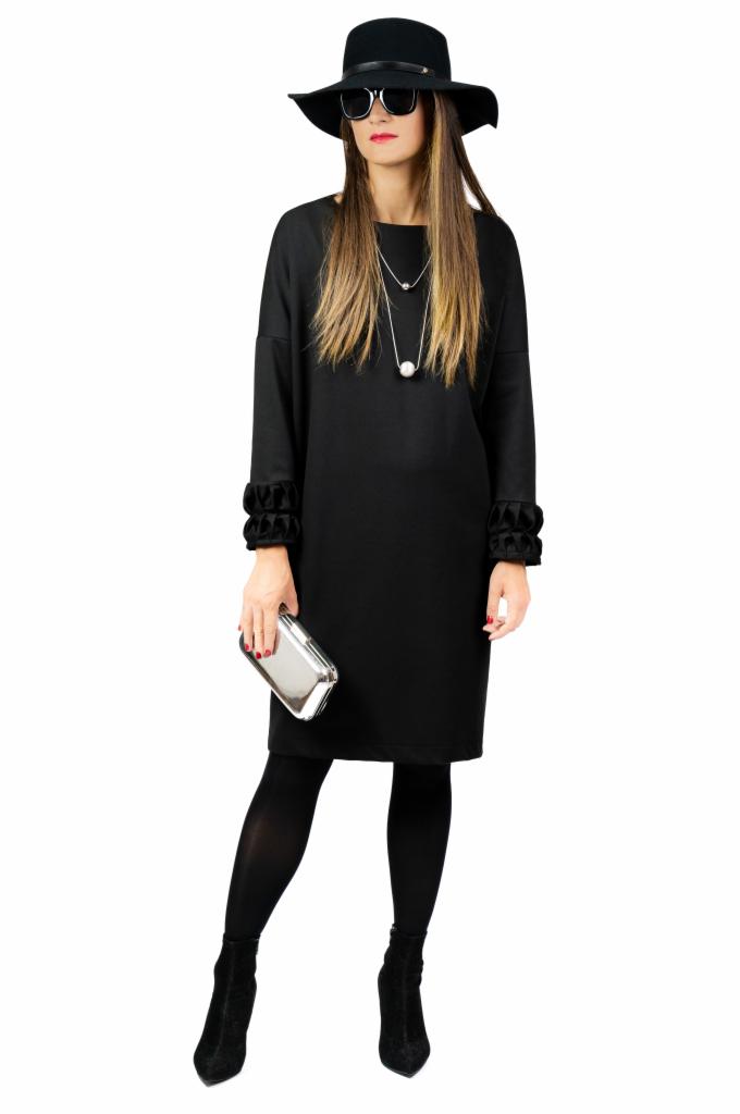 Kobieta w czarnej sukience, kapeluszu i okularach przeciwsłonecznych, trzymająca srebrną kopertówkę, na białym tle.