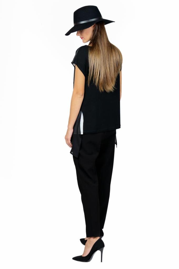 Kobieta z długimi blond włosami, ubrana w czarny kapelusz, bluzkę z asymetrycznym wykończeniem i czarne spodnie, pozuje tyłem na białym tle, prezentując elegancki, monochromatyczny strój.