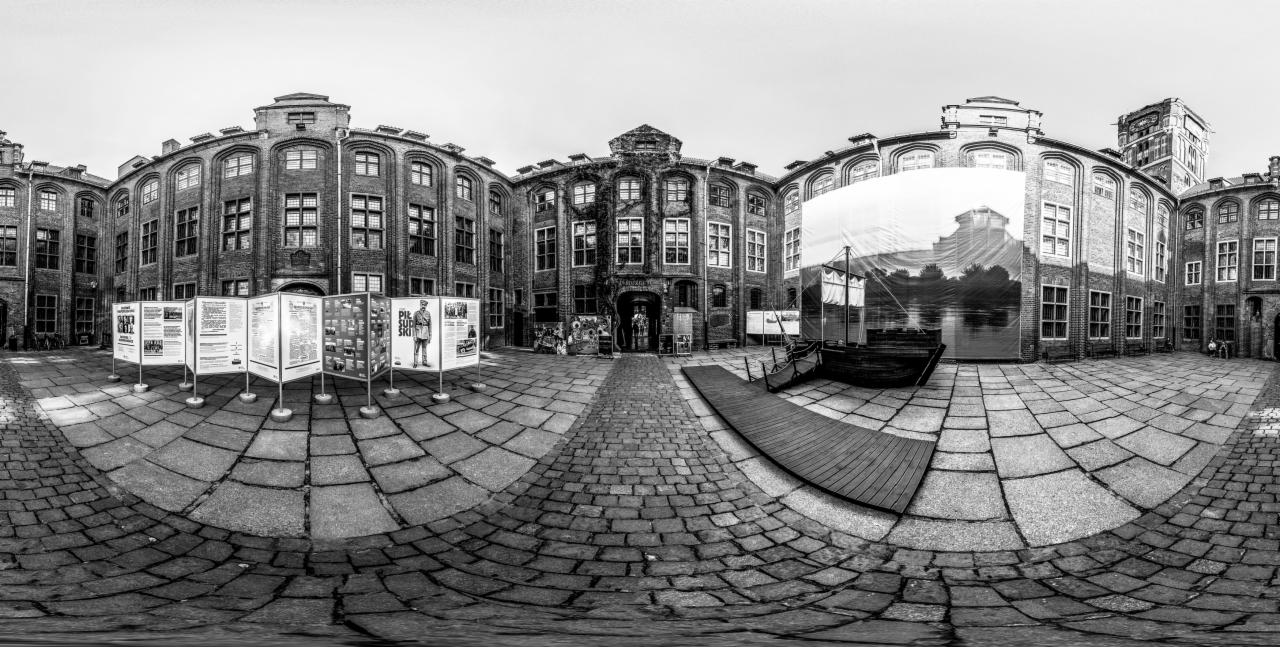Panoramiczne czarno-białe ujęcie dziedzińca z brukowaną nawierzchnią, otoczonego ceglanymi budynkami z rzędami okien, wystawą plansz informacyjnych i instalacją artystyczną przypominającą łódź...