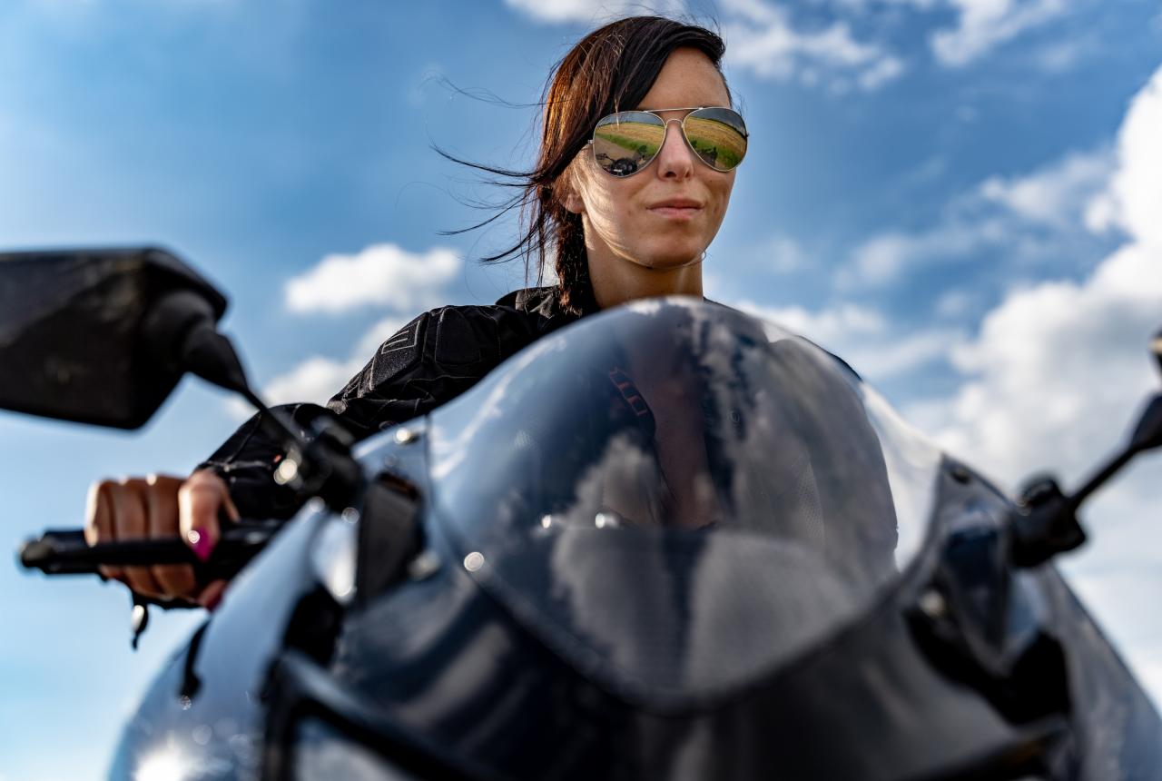 Kobieta w okularach typu aviator siedzi na motocyklu, w okularach odbija się krajobraz polny, w tle błękitne niebo z chmurami.