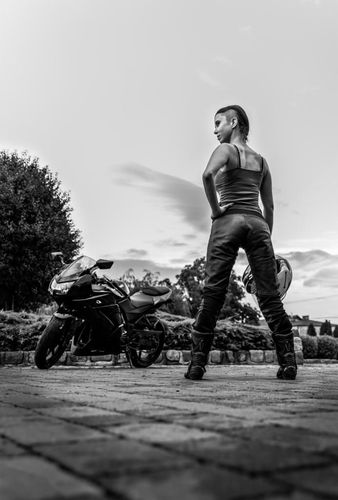 Czarno-białe zdjęcie kobiety w skórzanych spodniach i topie, stojącej tyłem z kaskiem w dłoni, obok czarnego motocykla na tle drzew i pochmurnego nieba.