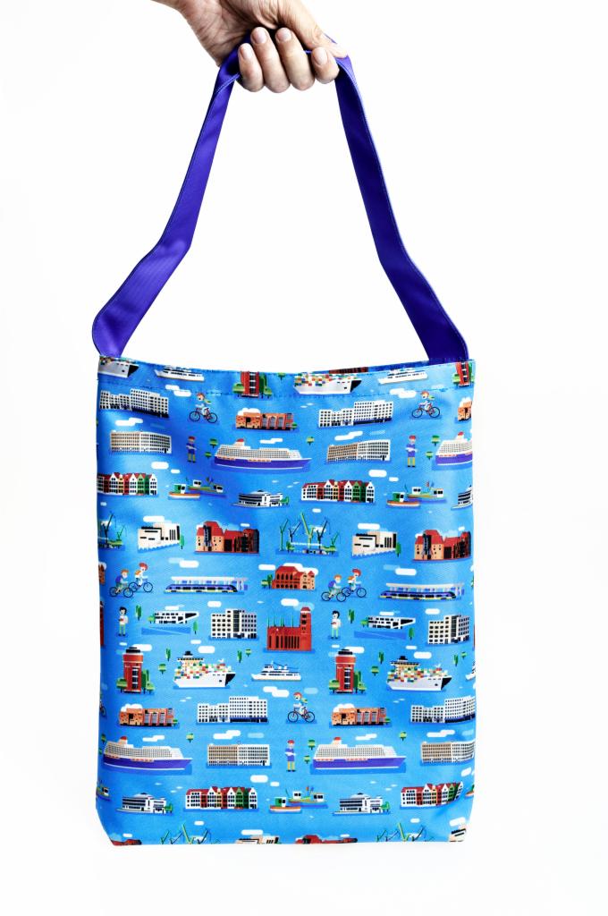Torba bawełniana z nadrukiem przedstawiającym budynki, statki i ludzi na rowerach, trzymana w dłoni, fioletowe uchwyty, jasne tło.
