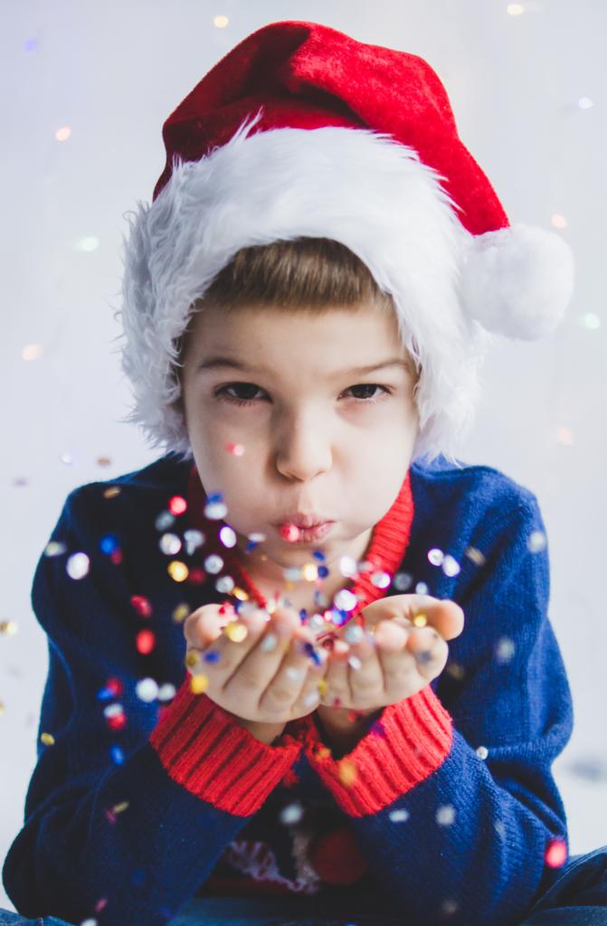 Chłopiec w czapce Świętego Mikołaja dmucha konfetti z dłoni na jasnym tle z delikatnymi światełkami.
