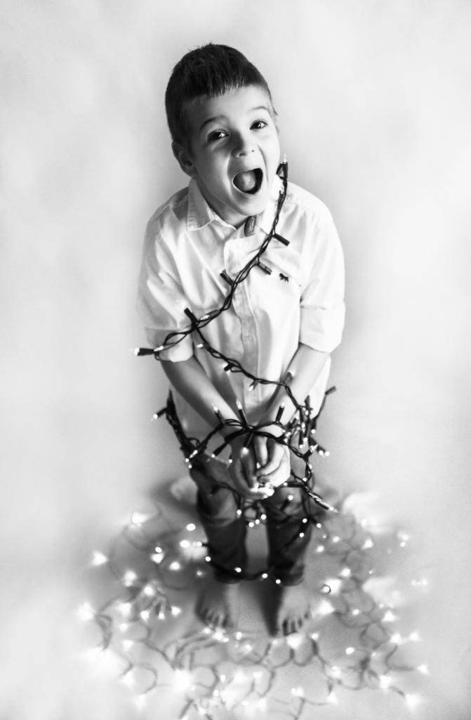 Czarno-białe zdjęcie chłopca z otwartymi ustami, owiniętego w światełka choinkowe, stojącego na podłodze z rozrzuconymi światełkami.