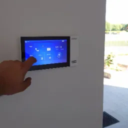 Palec wskazujący na ekranie dotykowym wideodomofonu Dahua zamontowanego na ścianie wewnątrz budynku, z widokiem na dom przez otwarte drzwi.