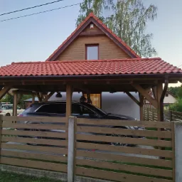 Zadaszone miejsce parkingowe z drewnianą konstrukcją i czerwoną dachówką, chroniące ciemny samochód osobowy widziane przez drewniany płot.