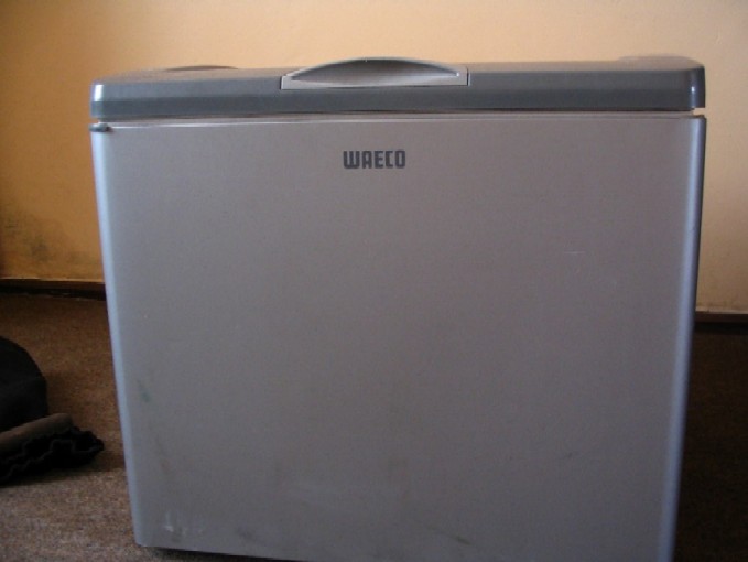 Szara lodówka turystyczna marki WAECO z uchwytem na górze, przeznaczona do użytku w ciężarówce, widoczna na tle ściany i dywanu.
