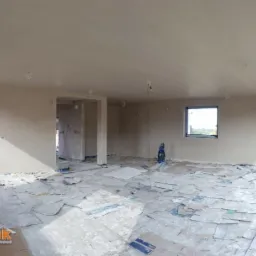 Wnętrze w trakcie prac wykończeniowych: świeżo położony jasny tynk na ścianach i suficie, podłoga zabezpieczona kartonem, widoczne okno oraz drzwi balkonowe, wystające kable oświetleniowe.