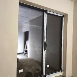 Świeżo otynkowana ściana wokół okna tarasowego z czarną ramą; widoczne ślady po pracach wykończeniowych i drabina w pomieszczeniu za oknem.