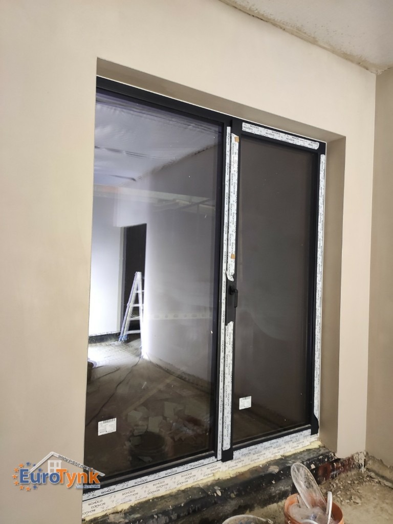 Świeżo otynkowana ściana wokół okna tarasowego z czarną ramą; widoczne ślady po pracach wykończeniowych i drabina w pomieszczeniu za oknem.
