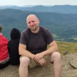 Mężczyzna z brodą siedzi na kamieniu na tle gór. Ubrany w szarą koszulkę i beżowe spodenki. W tle widoczne dzieci i torba. Krajobraz górski w tle.