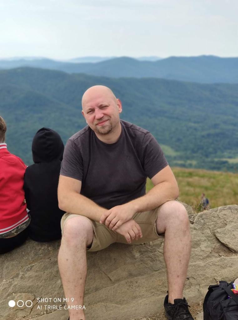 Mężczyzna z brodą siedzi na kamieniu na tle gór. Ubrany w szarą koszulkę i beżowe spodenki. W tle widoczne dzieci i torba. Krajobraz górski w tle.