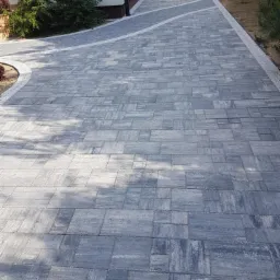 Szeroki, szary podjazd z kostki brukowej prowadzący do domu z garażem, z obrzeżem z drobnego kamienia i pasem z jasnej kostki, widok z góry.