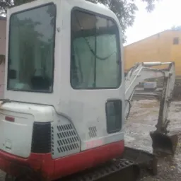 Niewielka, używana koparka na gąsienicach, z kabiną z widocznymi kroplami deszczu na szybach, stoi na błotnistym gruncie, z drzewami i budynkiem w tle.