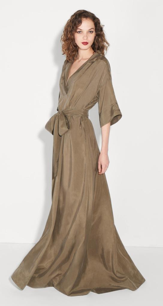 Modelka prezentuje długą, satynową sukienkę w odcieniu khaki z wiązaniem w talii, na białym tle.
