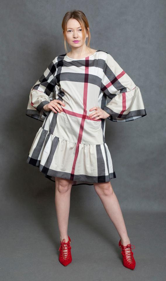 Kobieta pozuje w krótkiej sukience w kratę z falbaną i rozkloszowanymi rękawami, do tego czerwone sznurowane buty na obcasie na szarym tle.