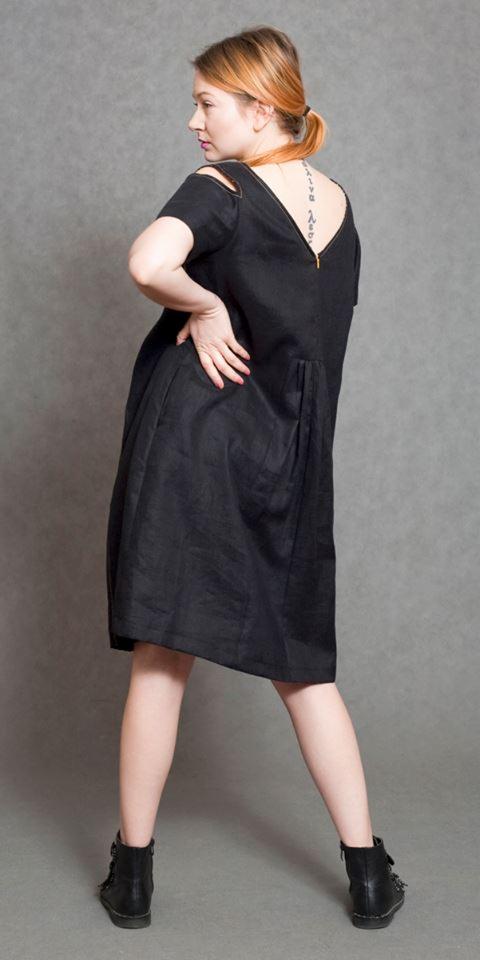 Kobieta pozuje w czarnej lnianej sukience z rozcięciami na ramionach i zamkiem błyskawicznym na plecach, widoczny tatuaż na karku i czarne botki na płaskiej podeszwie.