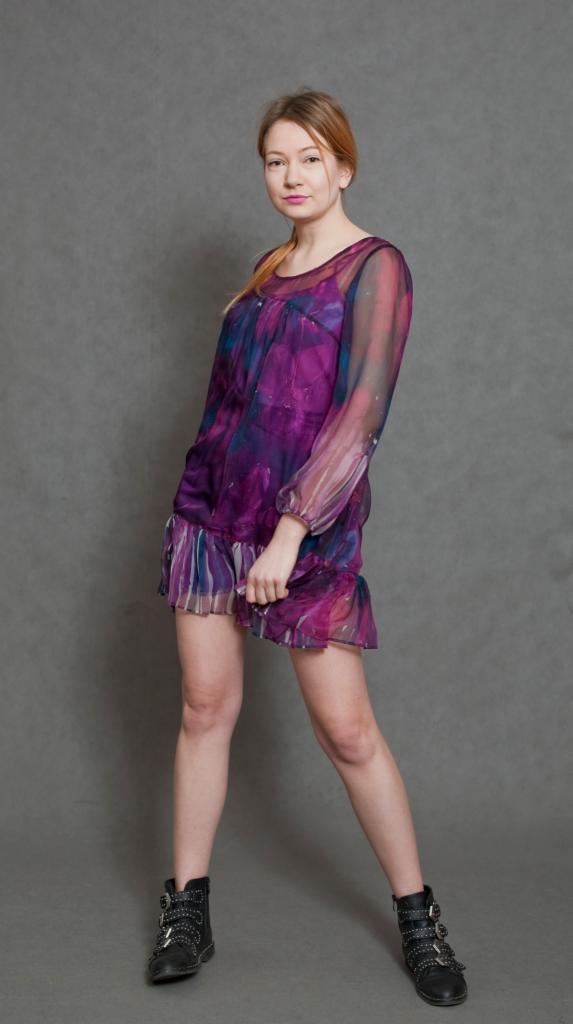 Kobieta w zwiewnej, fioletowej sukience z długimi rękawami i czarnych botkach na sznurowadła, pozująca na szarym tle.