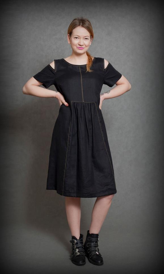 Kobieta w czarnej sukience z wycięciami na ramionach i ozdobnymi przeszyciami, pozująca na szarym tle w botkach z klamrami.