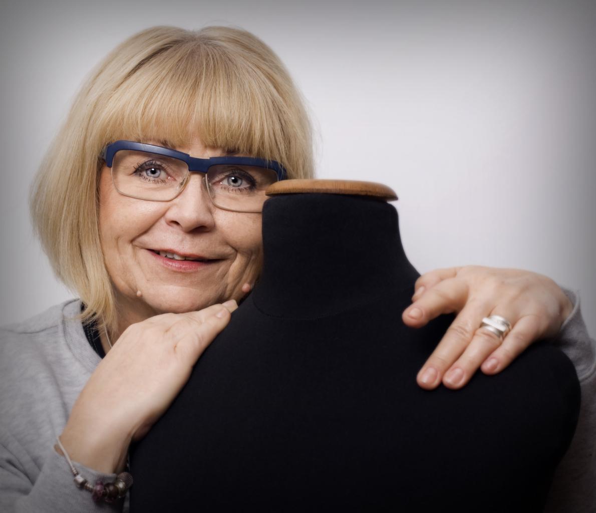 Uśmiechnięta blondynka w okularach z niebieskimi oprawkami, opierająca się o czarny manekin krawiecki z drewnianą szyjką, pierścionek na palcu.