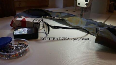 Stanowisko pracy projektanta odzieży: fragment tkaniny w kolorze khaki pod maszyną do szycia, pudełko szpilek z niebieskim wieczkiem, czarne okulary i pudełko z akcesoriami na białym stole.