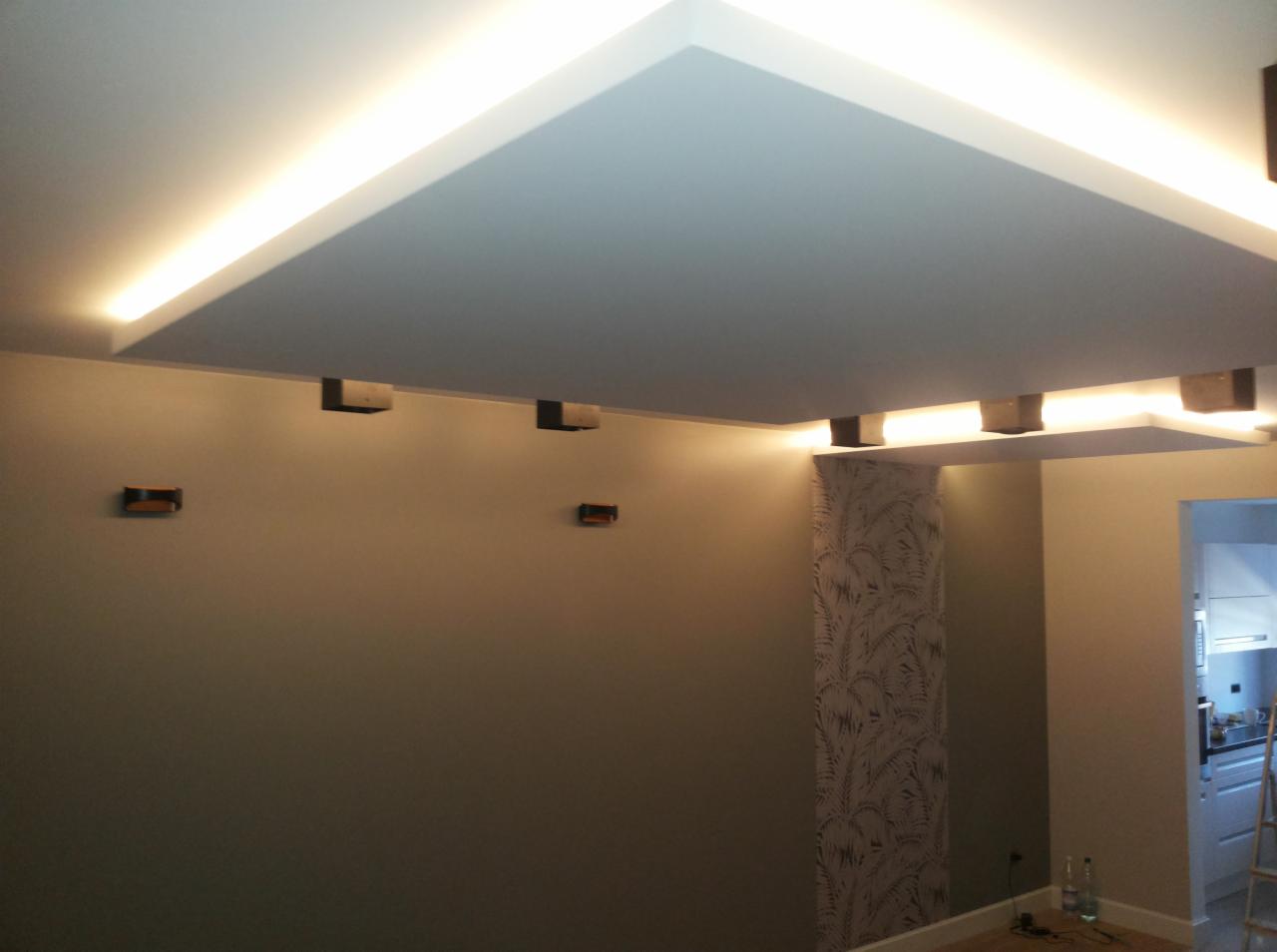 Nowoczesny sufit podwieszany z oświetleniem LED w ciepłej barwie, dekoracyjne wsporniki na ścianie, widoczny fragment wzorzystej tapety i kuchni w tle.