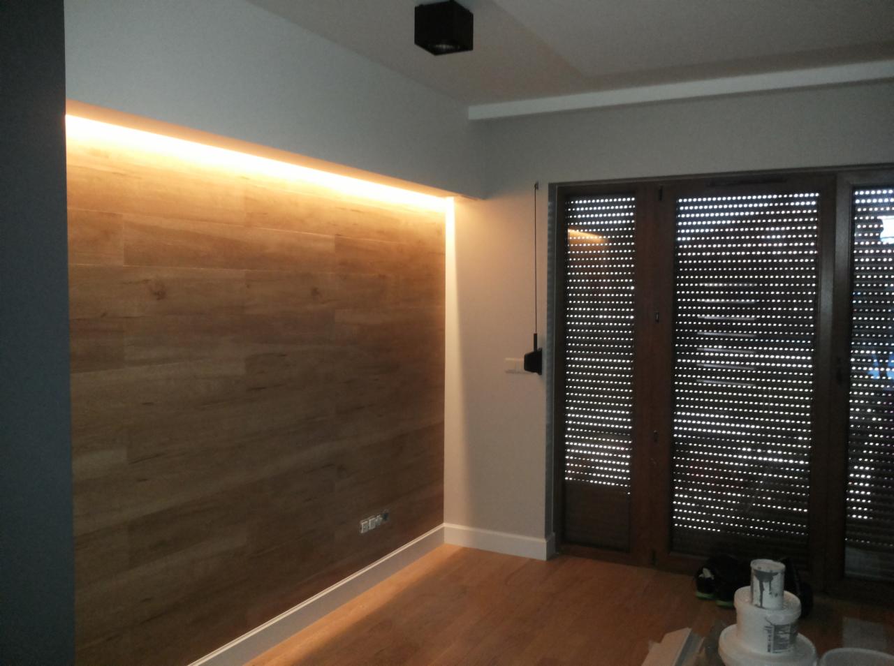 Drewniana ściana z podświetleniem LED wzdłuż górnej krawędzi, widoczne rolety zewnętrzne na oknach i podłoga z paneli, fragment wyposażenia malarskiego na podłodze.