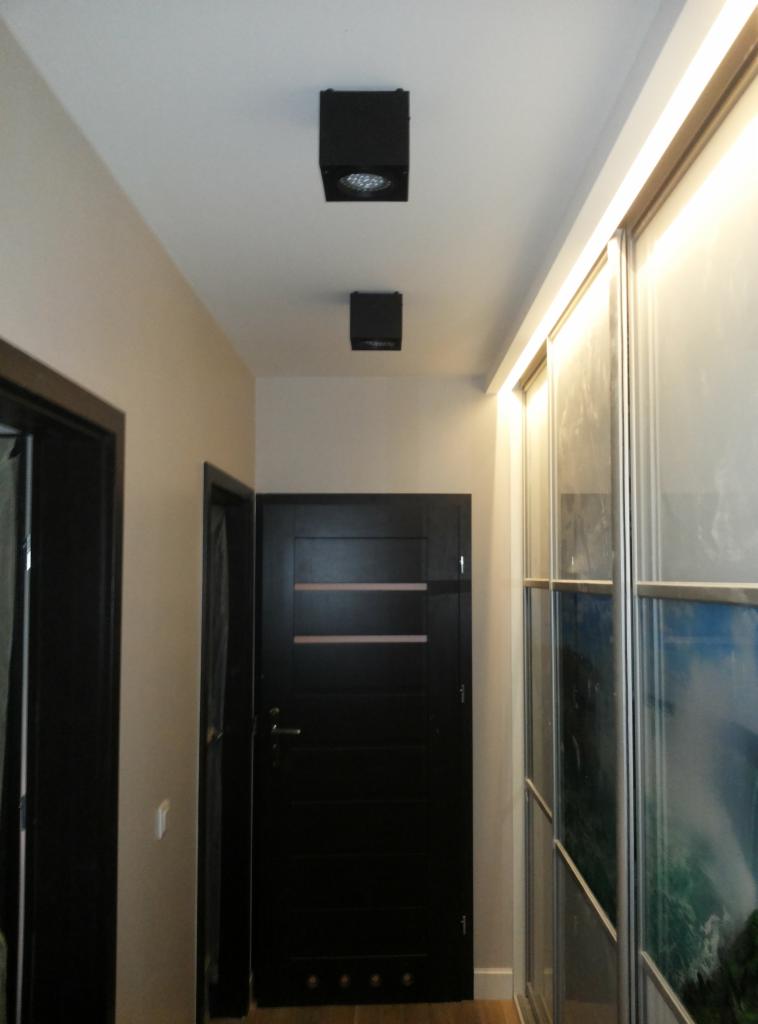 Korytarz z czarnymi, kwadratowymi lampami sufitowymi LED, drzwiami w ciemnym kolorze i przesuwną szafą z oświetleniem LED, w jasnej tonacji kolorystycznej.