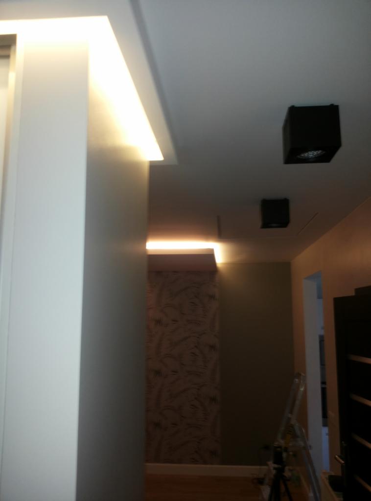 Korytarz z oświetleniem LED w suficie podwieszanym i czarnymi lampami sufitowymi, widoczna ściana z tapetą o wzorze roślinnym i drabina malarska w tle.