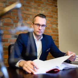 Mężczyzna w garniturze siedzi za biurkiem, przeglądając otwartą książkę lub dokumenty; w tle ściana z czerwonej cegły.