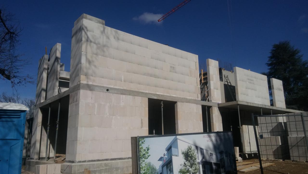 Wznoszenie ścian z bloczków betonu komórkowego na placu budowy, widoczny dźwig w tle i przenośna toaleta z boku.