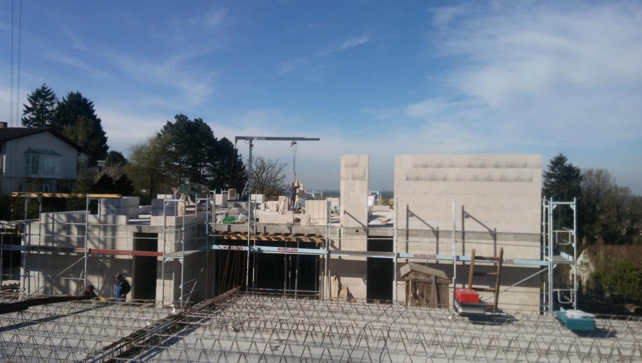 Budowa domu z pustaków: widok na wznoszone ściany, rusztowania, stalowe zbrojenie fundamentów oraz dźwig na placu budowy w słoneczny dzień.