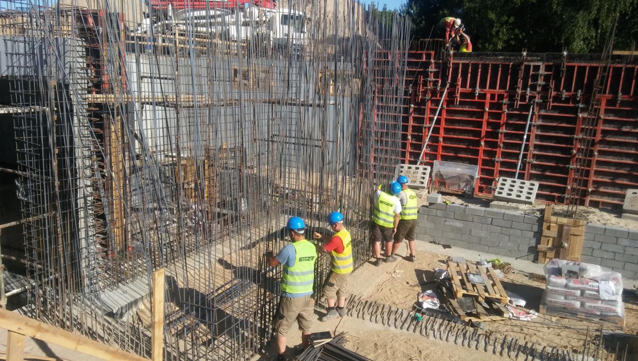 Ekipa budowlana w kaskach i kamizelkach odblaskowych pracuje przy zbrojeniu fundamentów pod budynek, widoczne szalunki i materiały budowlane.