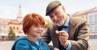 Uśmiechnięty chłopiec z rudymi włosami i starszy mężczyzna w tweedowej marynarce i kaszkiecie, trzymający monetę, na tle Rynku w Rawie Mazowieckiej. Scena sugeruje rozmowę o oszczędzaniu lub pożyczce.