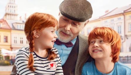 Uśmiechnięty dziadek w kaszkiecie i muszce otoczony dwójką rudowłosych wnucząt na tle miejskiego rynku z zabytkową architekturą.