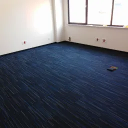 Pokój biurowy z nową, niebieską wykładziną dywanową w paski, widoczne gniazdka elektryczne na ścianach i otwór rewizyjny w podłodze.