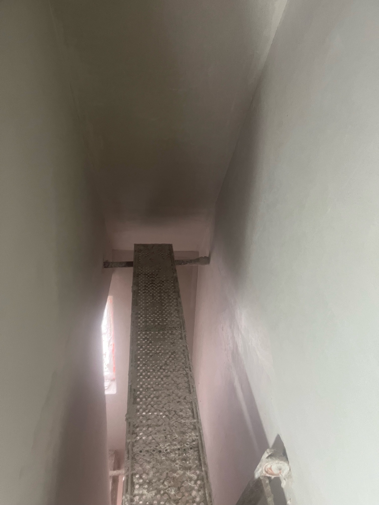 Wnętrze w trakcie prac tynkarskich, widoczne ściany pokryte świeżym tynkiem oraz metalowy ruszt w wąskim pomieszczeniu, z widocznym oknem w tle.