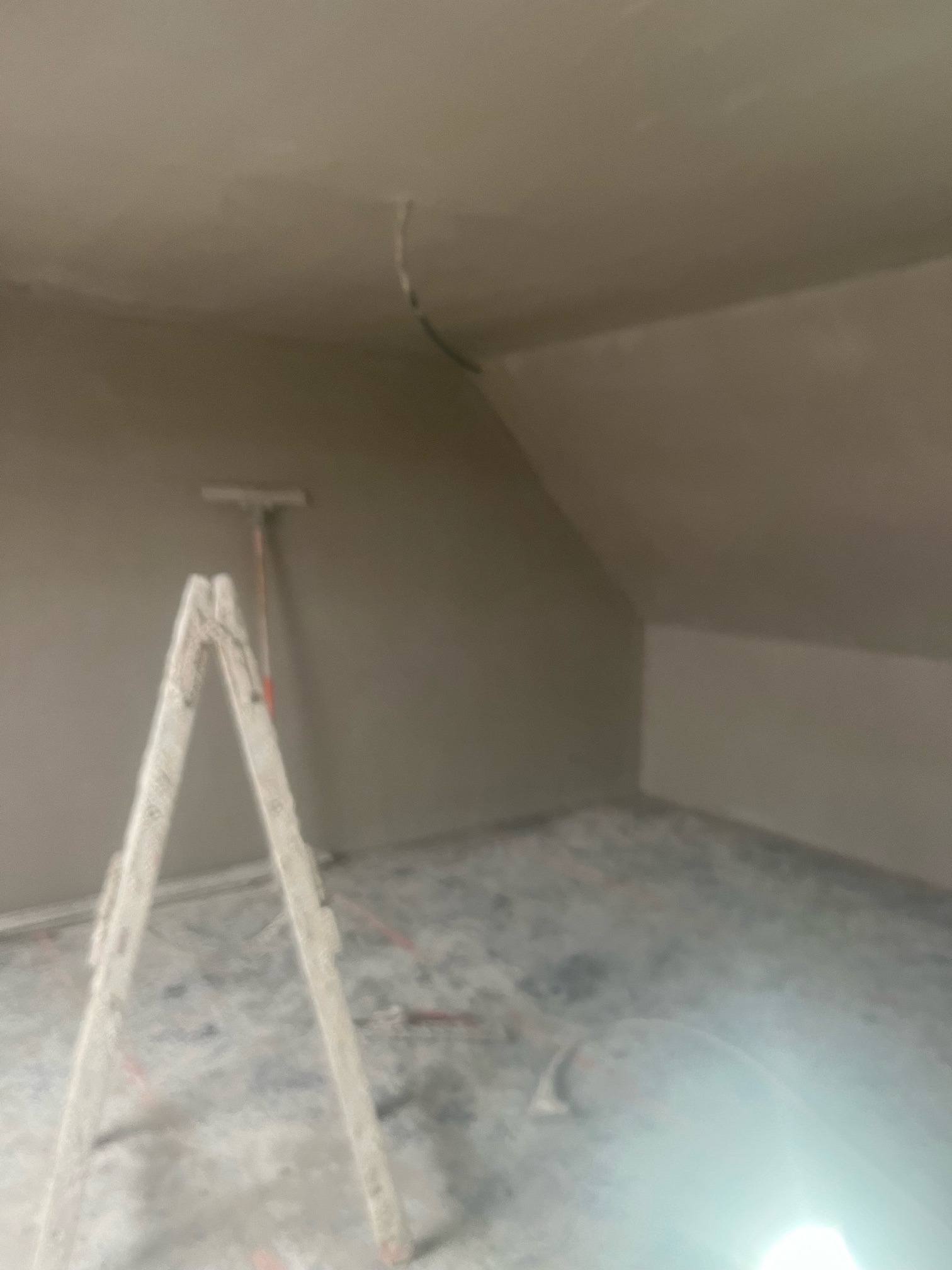 Pokój na poddaszu po nałożeniu tynku, z widoczną drabiną i narzędziem do rozprowadzania tynku przy ścianie.