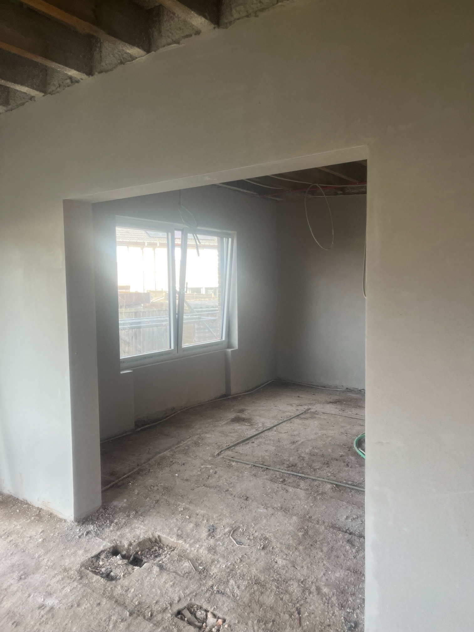 Wnętrze pokoju w trakcie remontu, świeżo otynkowane ściany w jasnym odcieniu szarości, widoczne okno z widokiem na zewnątrz, surowy betonowy strop i niezagospodarowana podłoga z wystającymi...
