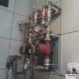 Rozdzielacz centralnego ogrzewania z pompą obiegową zamontowany na ścianie w łazience w Krakowie. Widoczne liczne rury, zawory i przyłącza. Tło stanowią białe płytki ceramiczne.