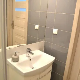 Umywalka z szafką w łazience, szare płytki na ścianie, lustro z odbiciem drzwi z przeszkleniami.